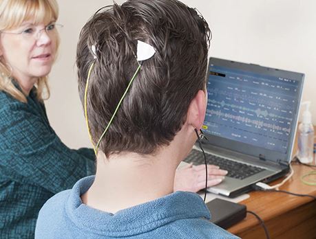 A male sits with electrodes attached to back of head and a screen showing his brain activity
