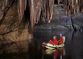 Two males geologists in a raft paddle through a cave with stalactites hanging just above them