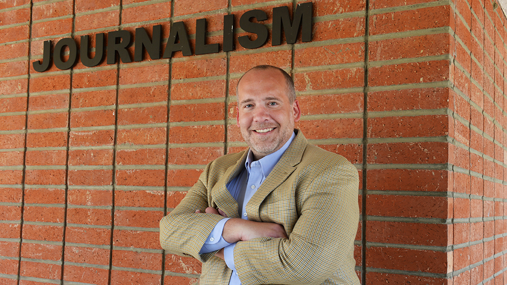 photo of jeremy shermak in front of brick building with letters spelling journalism behind him