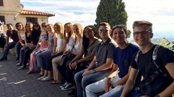 Group of male and female students smiling sitting on a short wall overlooking hills of Italy